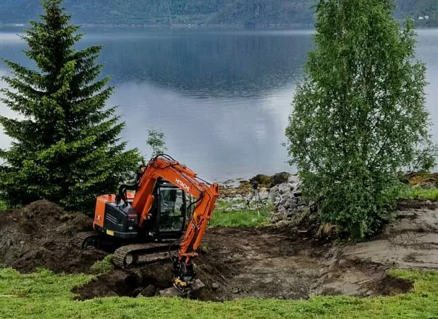 Gravemaskin utfører grunnarbeid ved innsjø, omgitt av trær og natur i landlige omgivelser