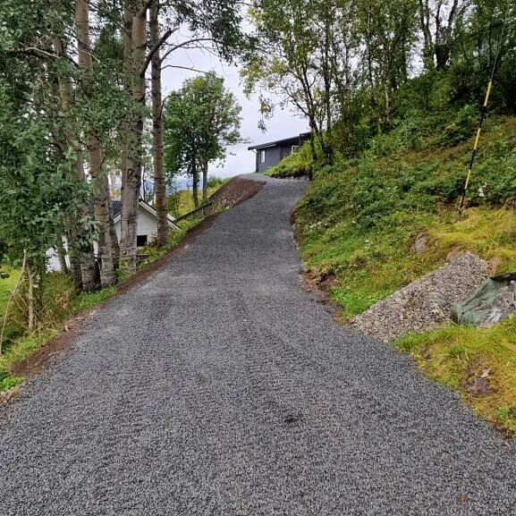 Nygruset innkjørsel som slynger seg oppover mot bolig, omgitt av trær og grønt terreng